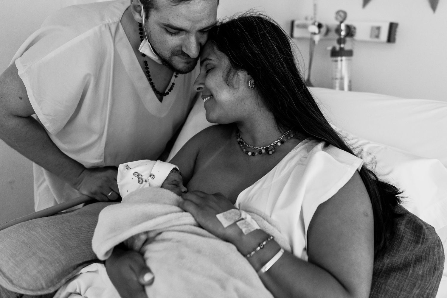 madre en cama se sanatorio dando la teta a su bebé recién nacido con su cabeza reclinada sobre la de su marido parado