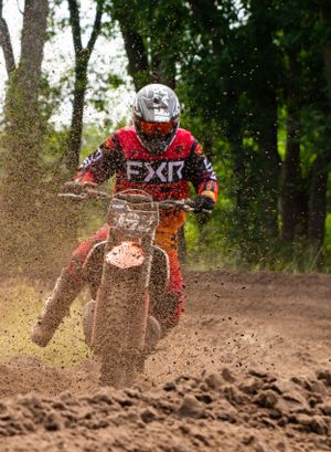 Dirt bike rider carves through deep track ruts while spraying mud on a wooded motocross course.
