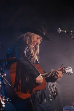 Musician in fringed jacket playing acoustic guitar on dimly lit stage during performance.