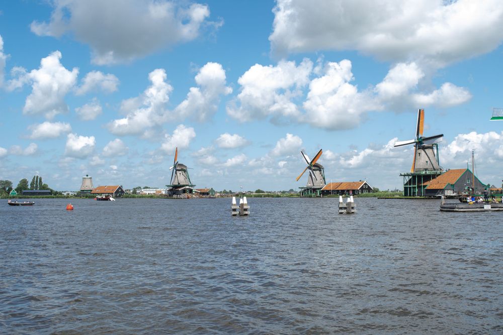 Traditional Dutch windmills stand along a waterway with boats sailing under blue skies and puffy clouds.