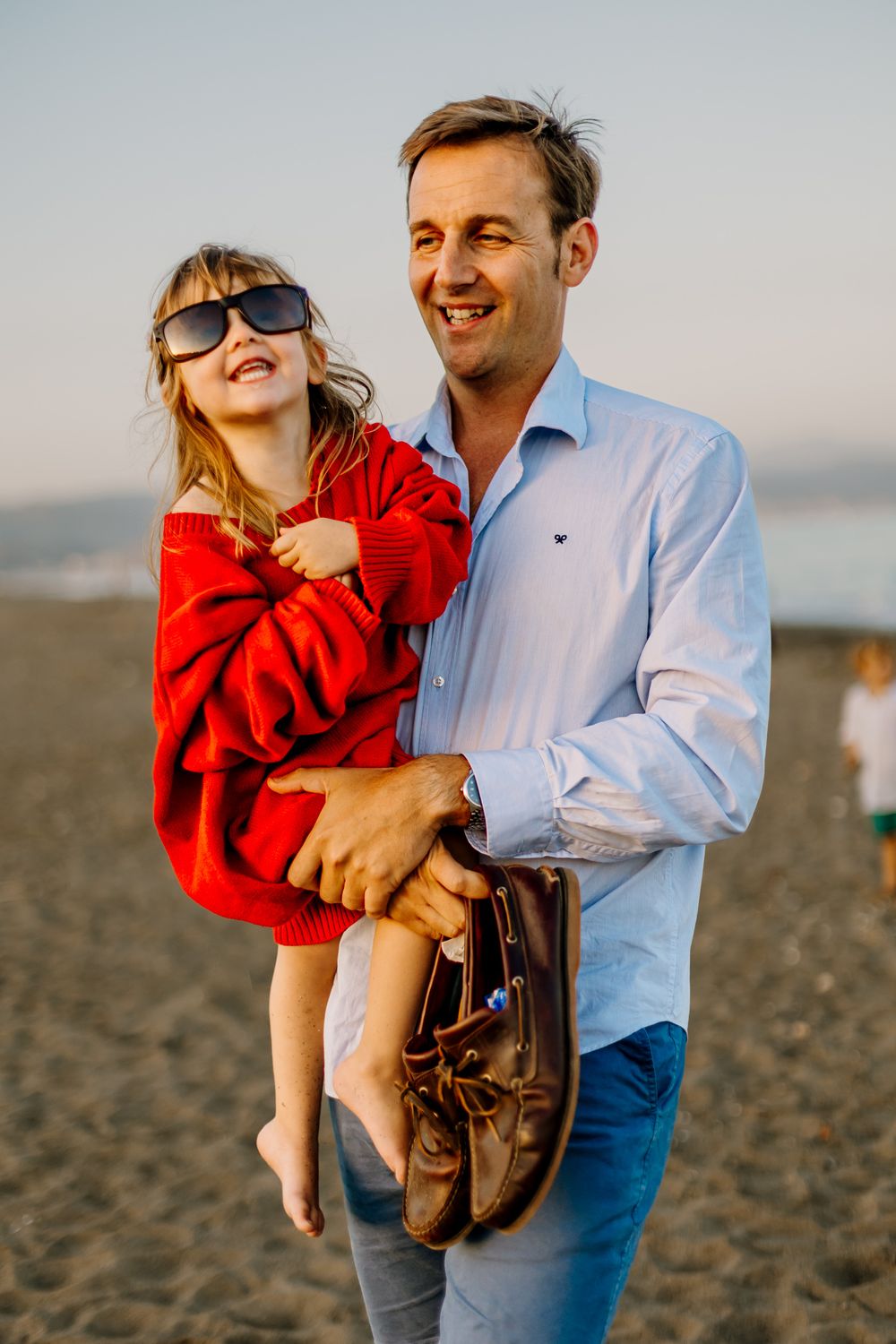 Father holds his daughter in his arms, while she happily wears his red sweater and sunglasses.