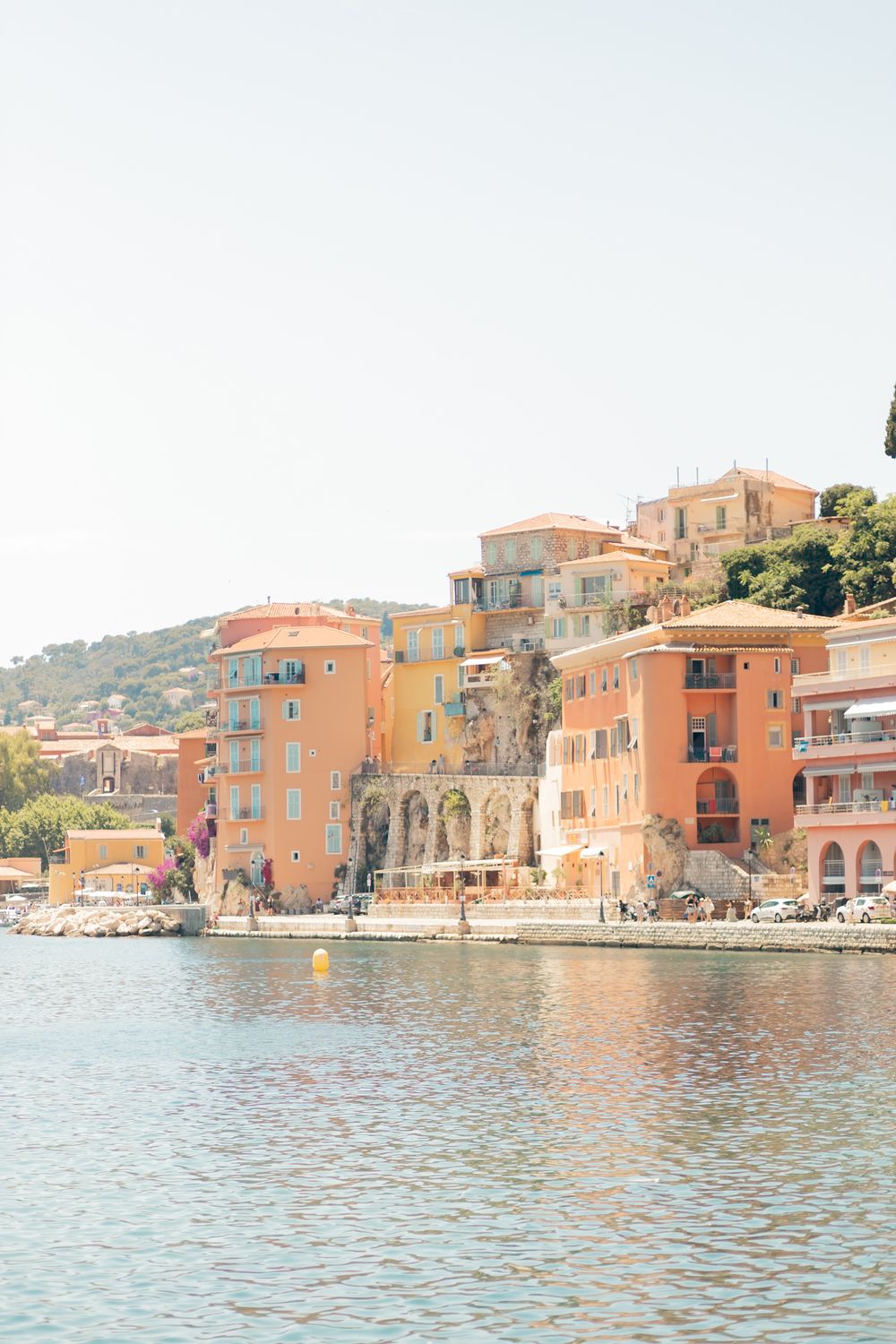 Colorful Mediterranean waterfront buildings in warm orange and yellow tones overlooking calm blue waters on a sunny day.