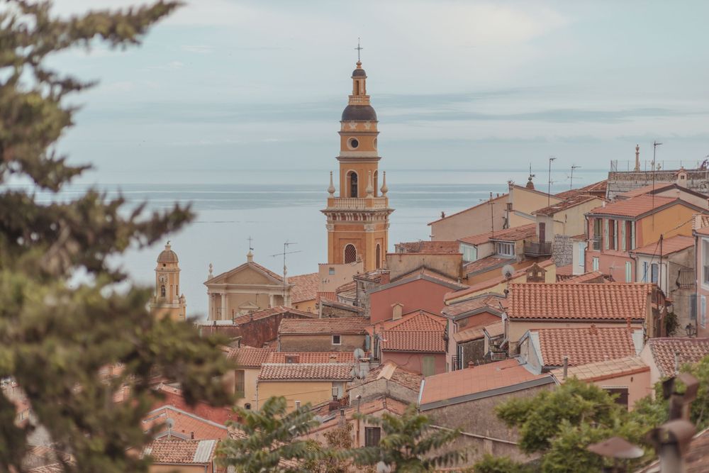 Aerial view of Menton old town rooftops with church bell tower and Mediterranean Sea in background.
