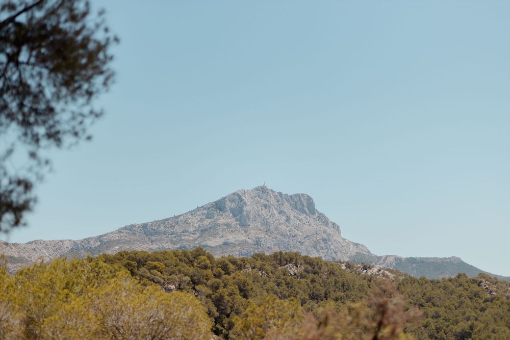 A rocky mountain peak rises above lush green trees under a clear blue sky in a serene Mediterranean landscape.