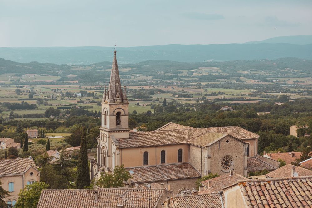 Aerial view of a historic stone church with a tall spire in a French village, surrounded by lush green valley landscape.