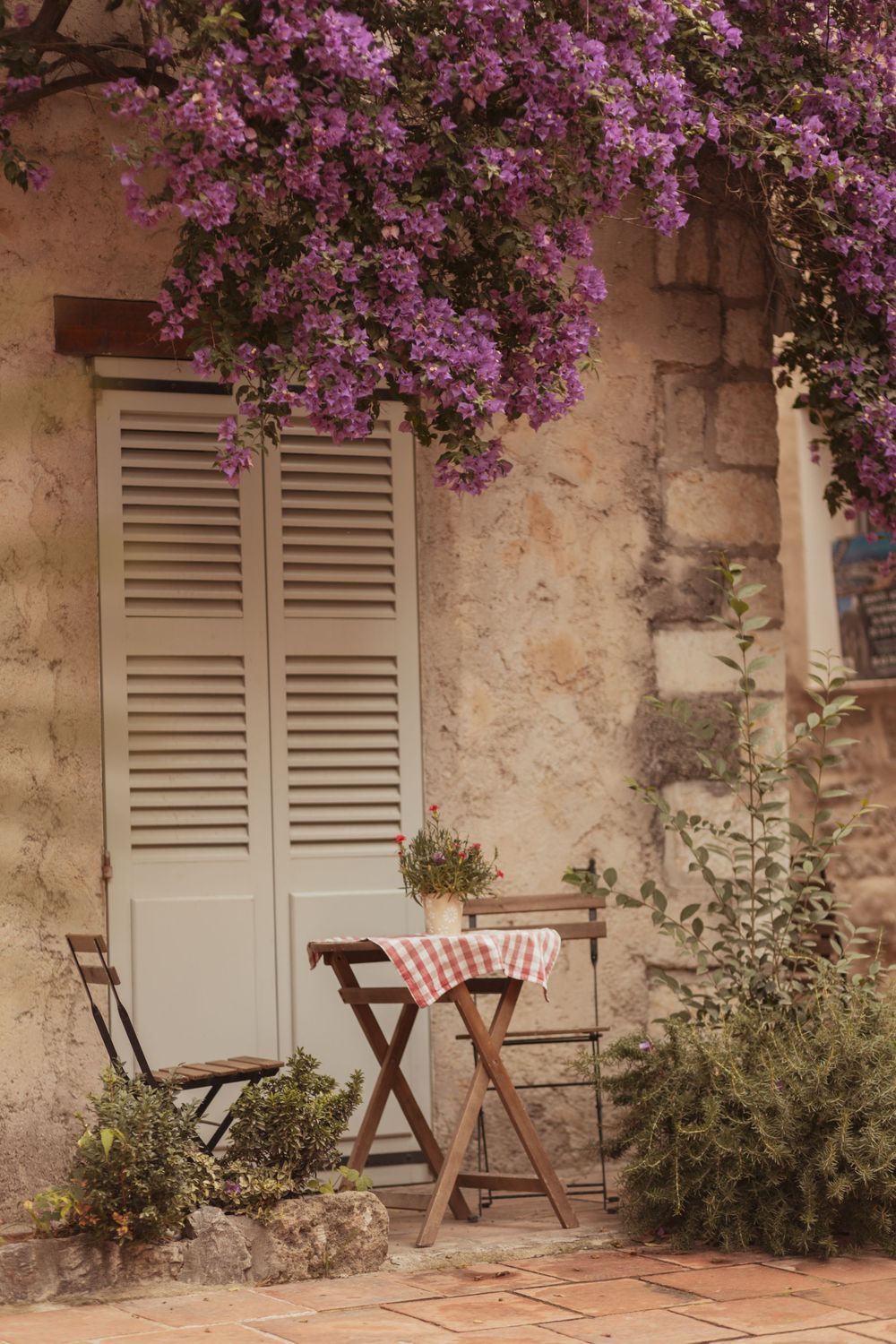 Charming French cafe terrace with bistro table, chairs, red checkered tablecloth, louvered door, and purple bougainvillea blooms.