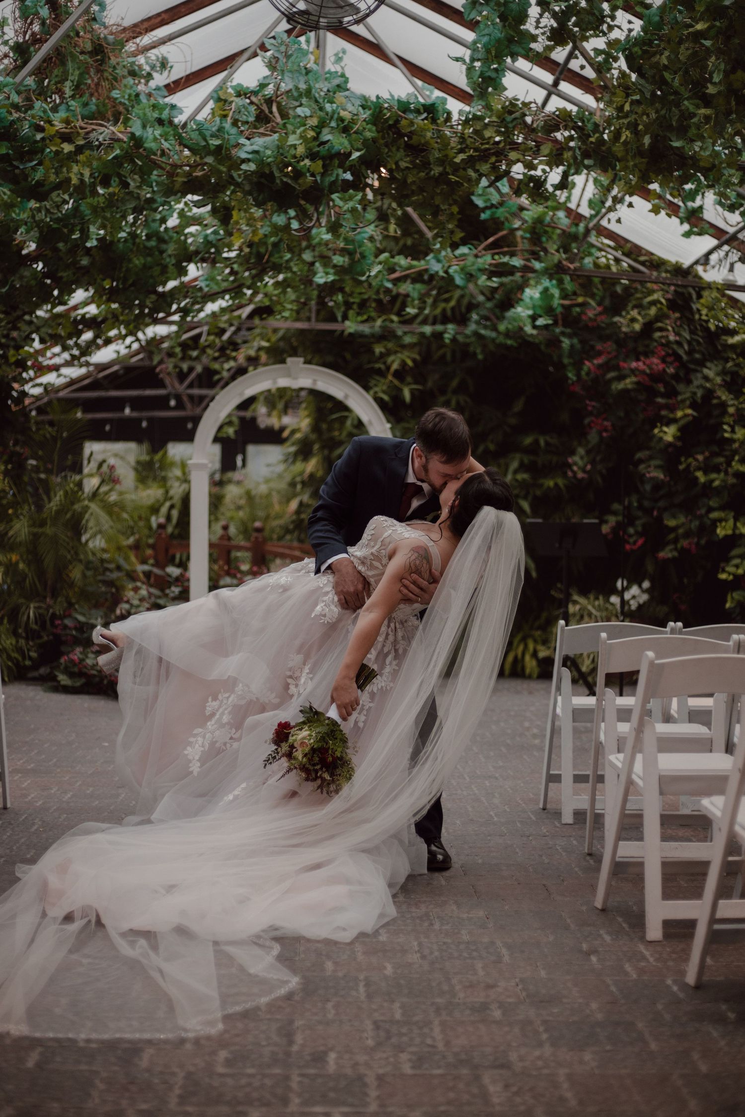 The groom dips the bride in the middle of the ceremony aisle to steal a kiss in a greenhouse event space.