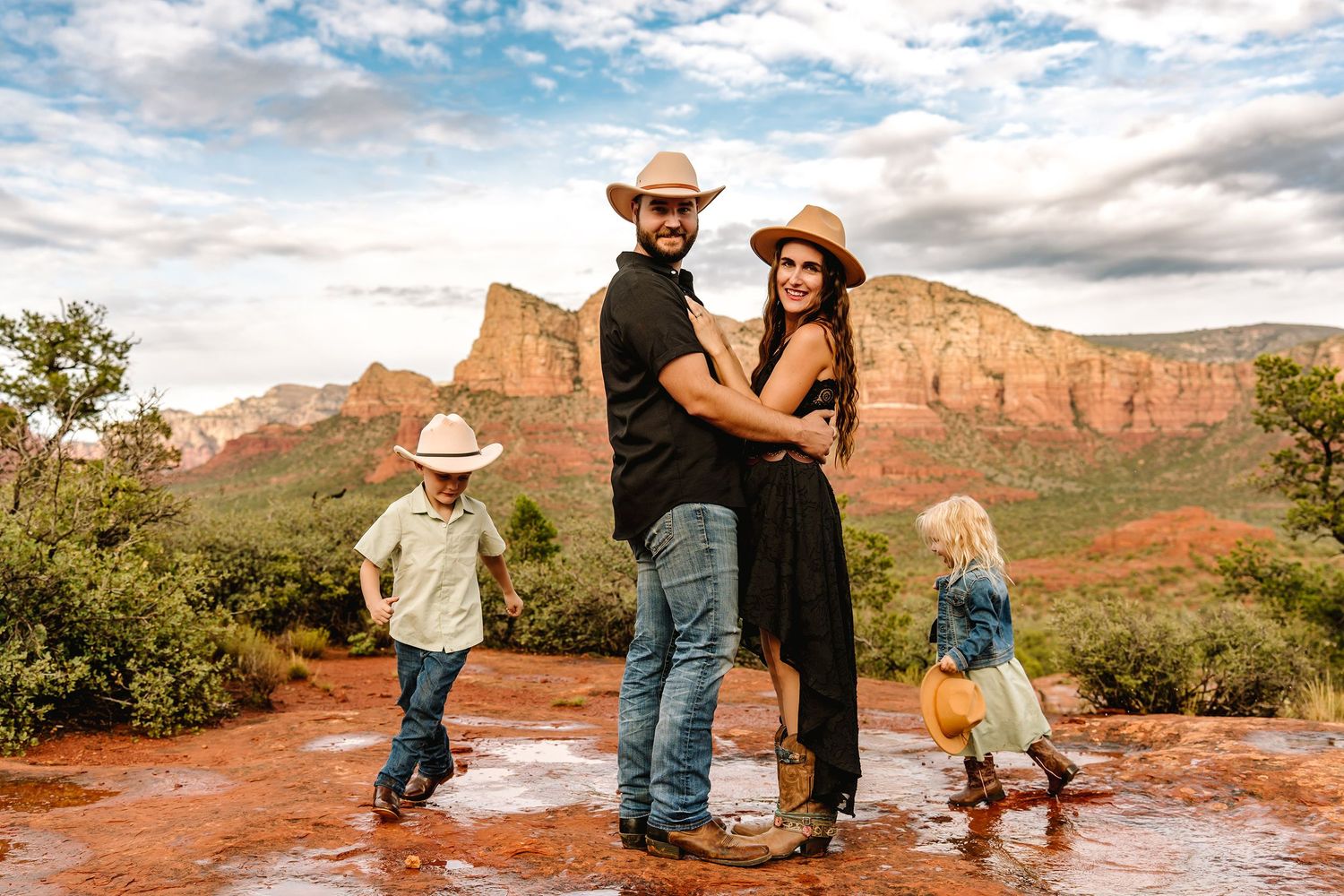 Family in cowboy hats poses on red rocks in Sedona, Arizona, with children playing nearby and red rock buttes in background.