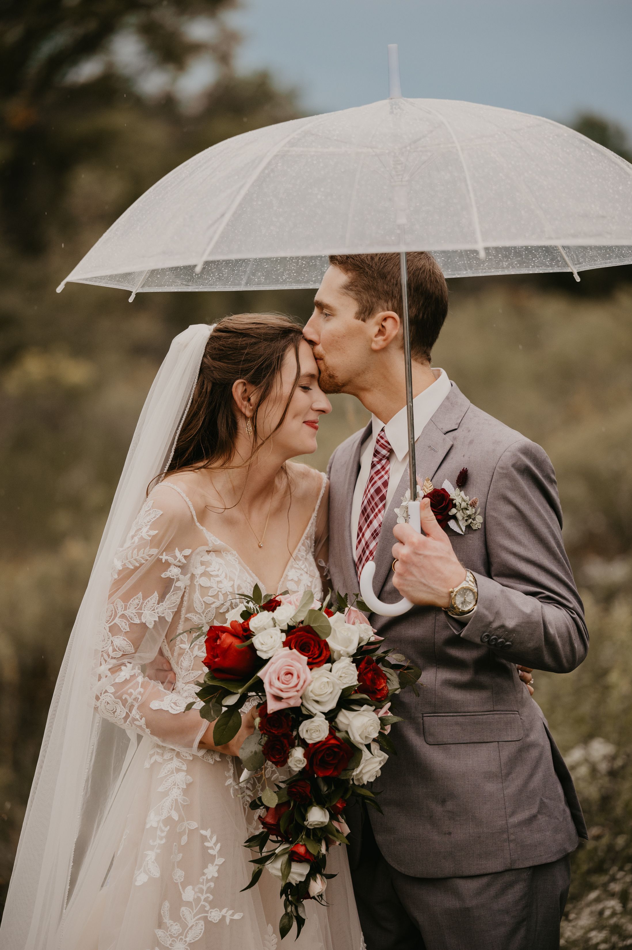 Bride and groom sharing a kiss under a clear umbrella on their rainy wedding day.