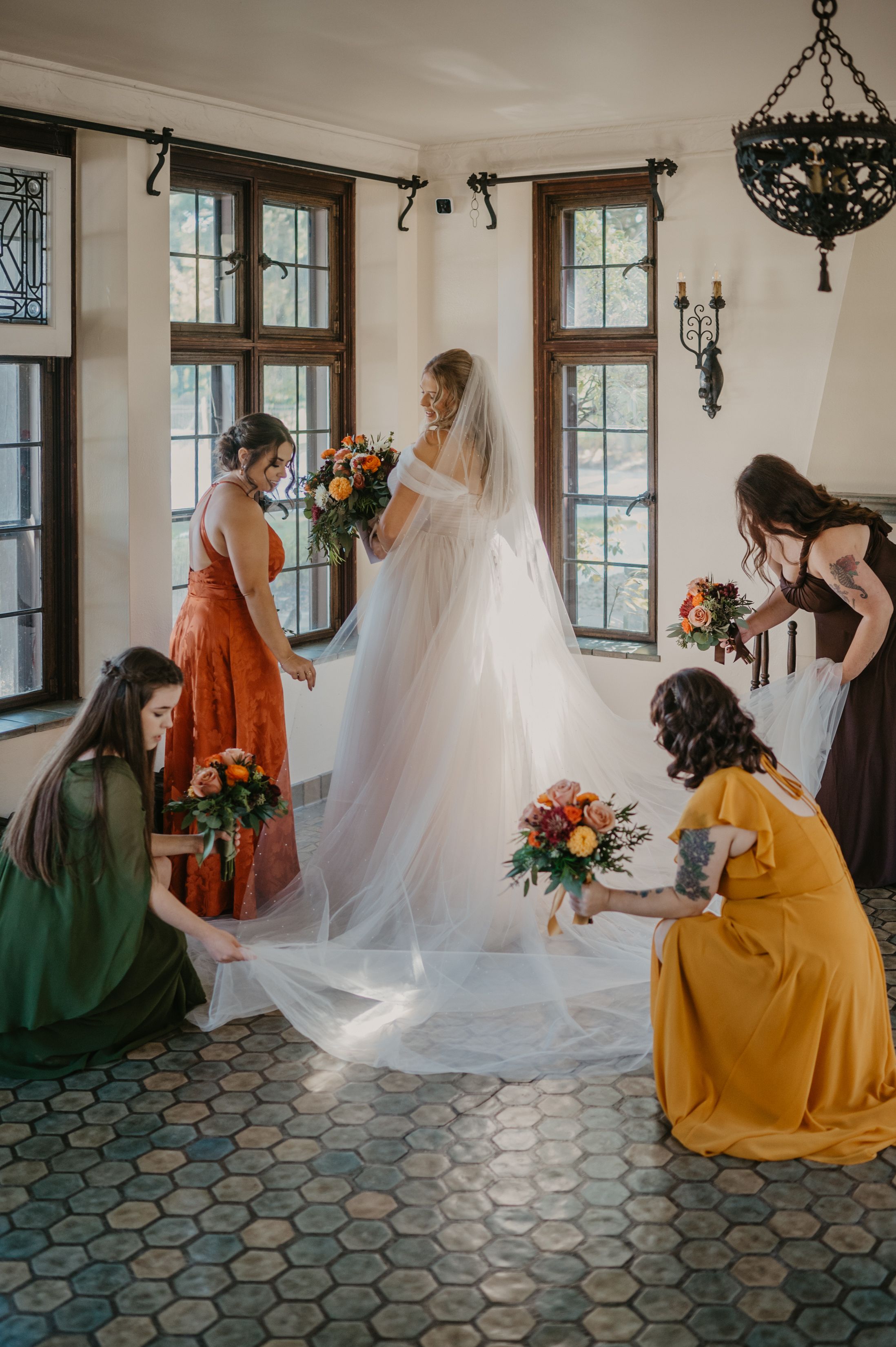 Bridesmaids in colorful dresses arranging bride's cathedral veil in elegant windowed room.