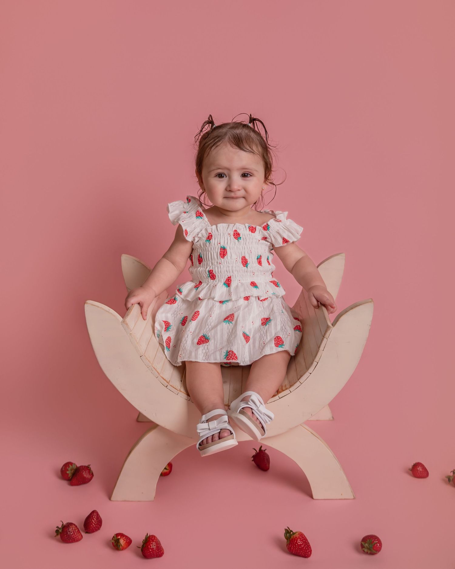 Toddler girl in strawberry print dress sitting on wooden chair with pink background and fresh strawberries.