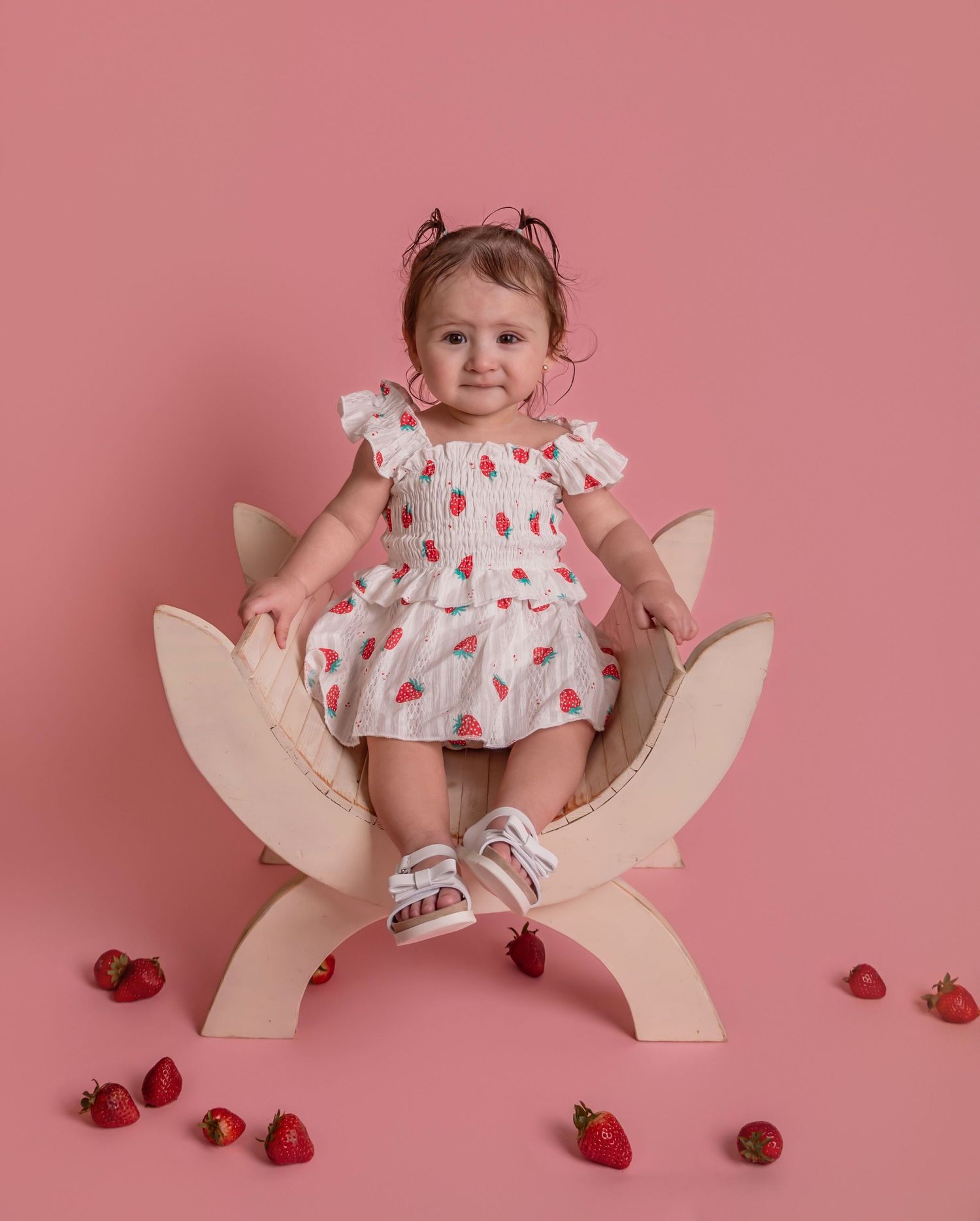 Baby girl in strawberry print dress sits on wooden flower chair with fresh strawberries on pink background.