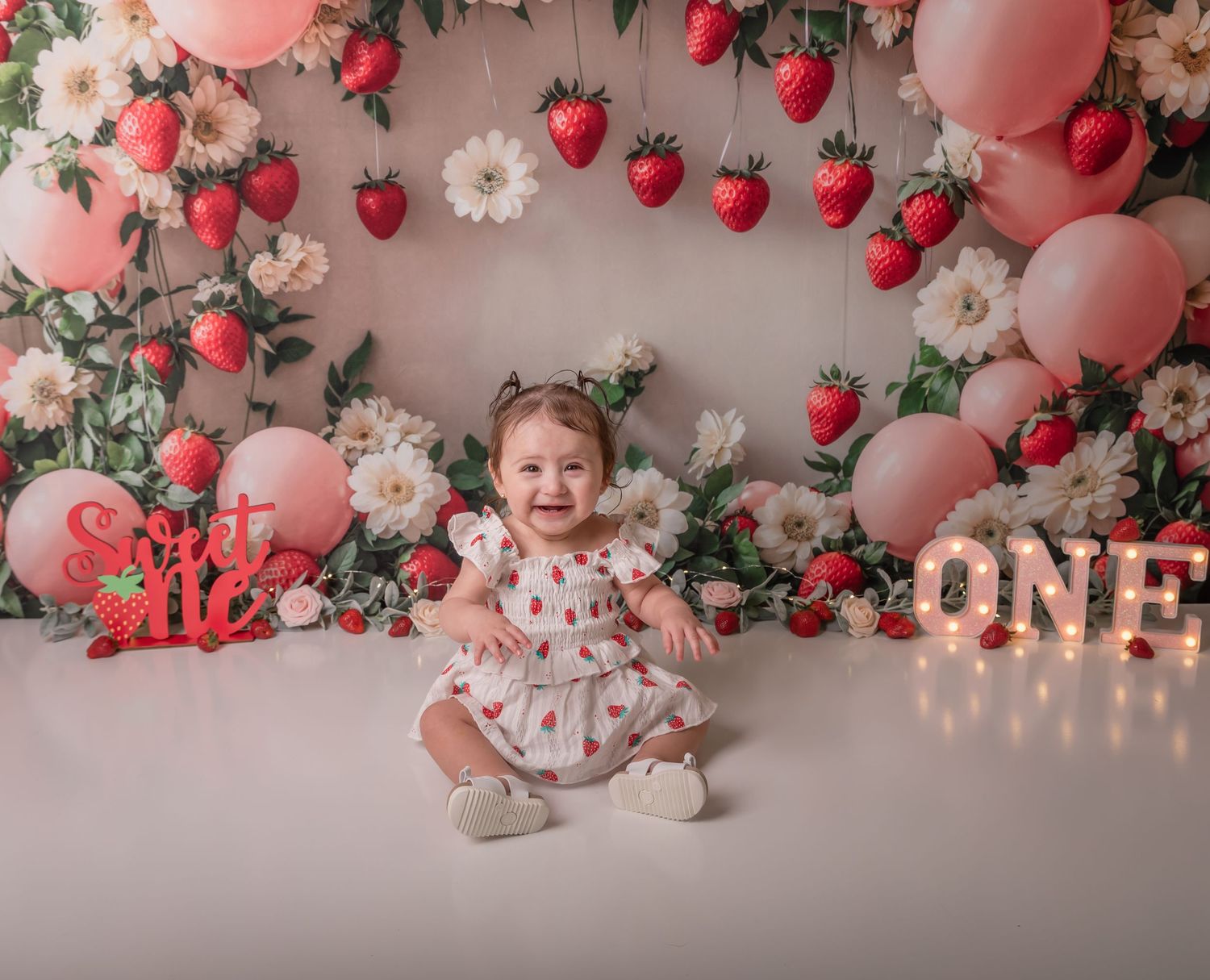 Smiling baby girl in strawberry dress sits before floral backdrop with pink balloons and ONE sign at first birthday cake smash.