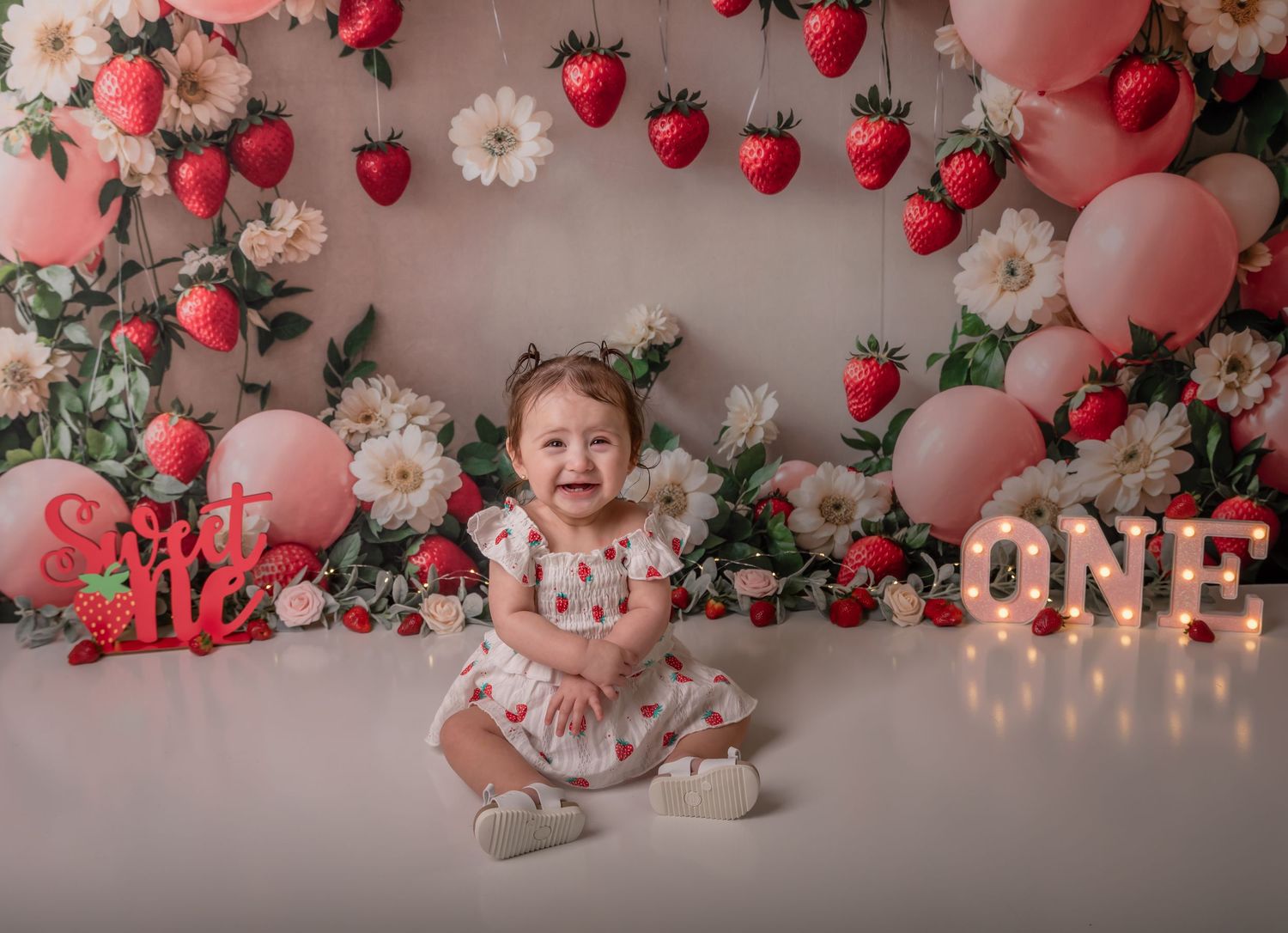 Smiling baby girl in strawberry dress sits before floral backdrop with balloons and ONE sign at first birthday cake smash.