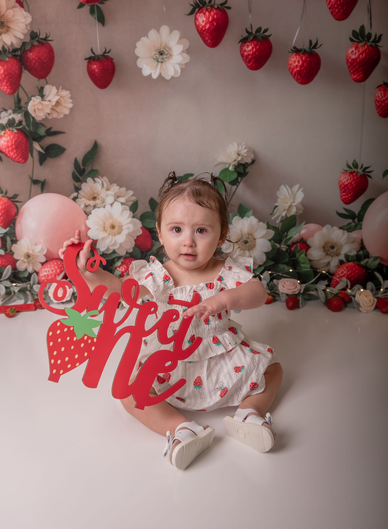 Baby girl in strawberry print dress sits before floral backdrop holding Sweet One sign at first birthday cake smash.