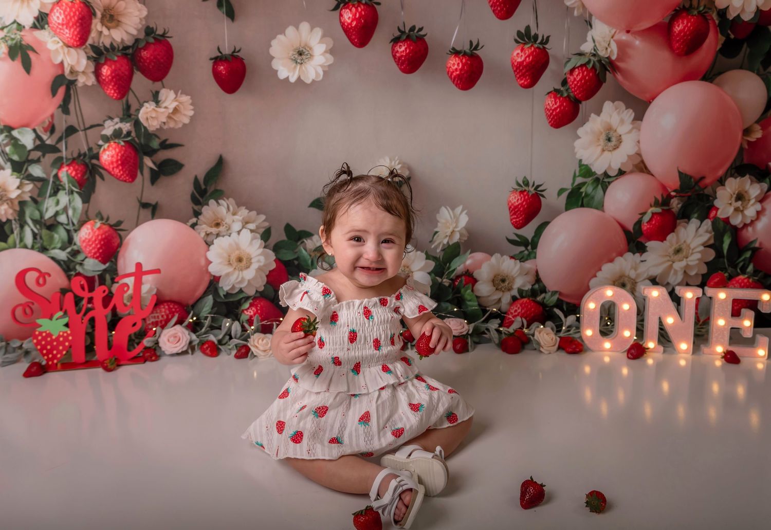Baby girl in strawberry dress sits smiling at first birthday cake smash with floral balloon backdrop.