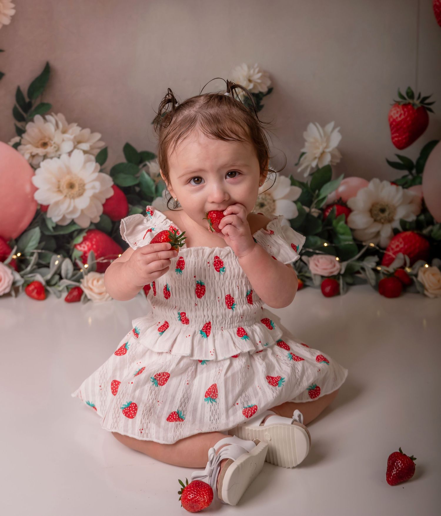 Adorable baby girl in strawberry print dress eating fresh strawberries during first birthday cake smash photo session.