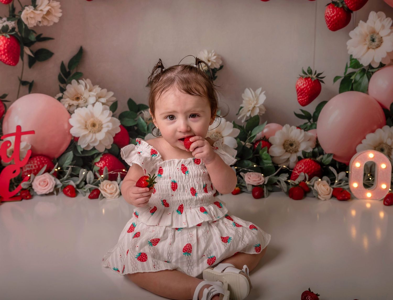 Baby girl in strawberry dress sits among floral backdrop with balloons and strawberries for first birthday cake smash.
