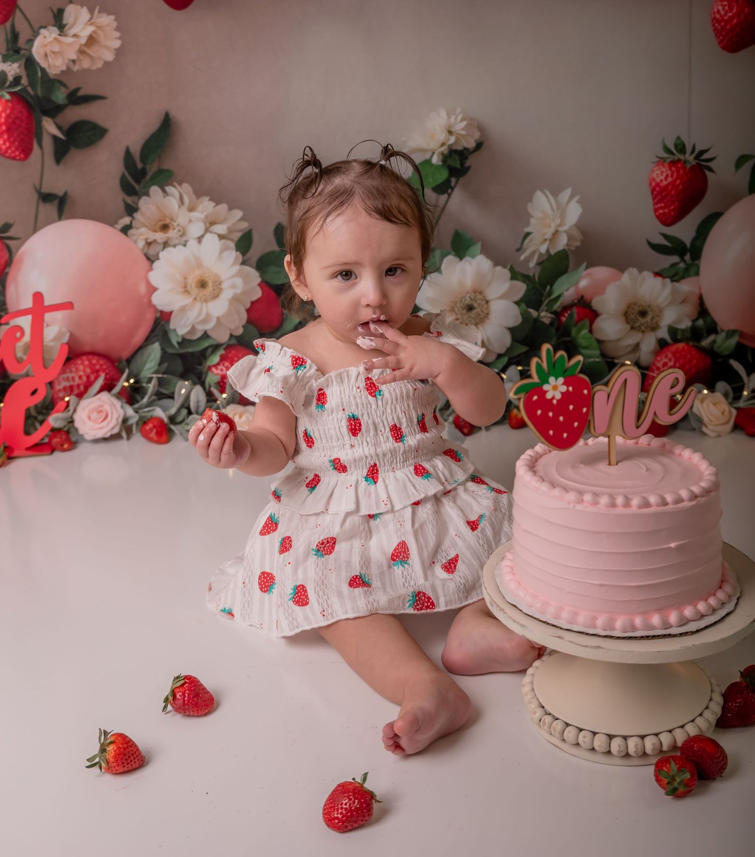 Baby girl in strawberry dress sits beside pink first birthday cake with floral backdrop and strawberry decorations.