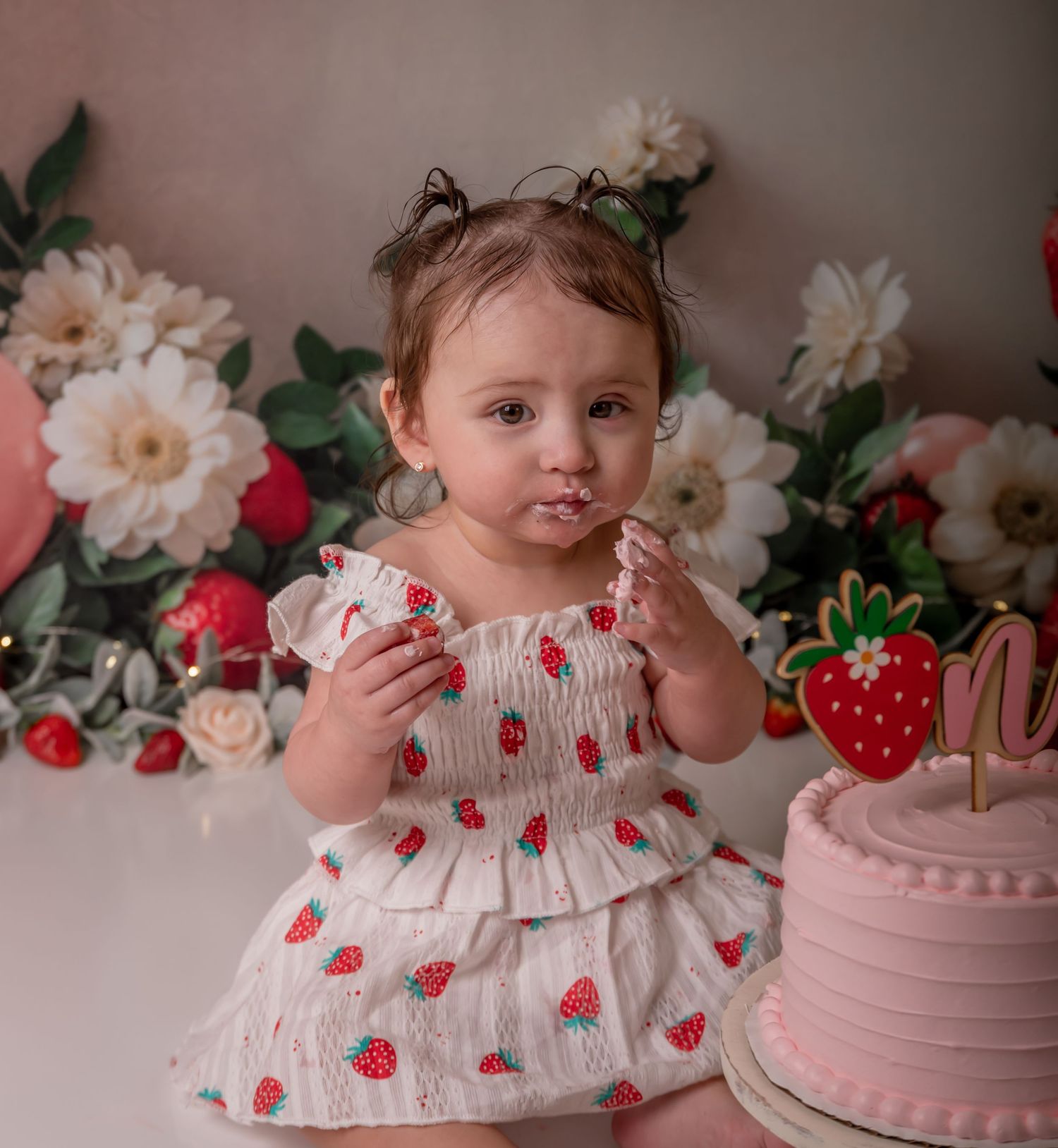 Toddler girl in strawberry dress eating cake at her first birthday party with pink cake and floral backdrop.