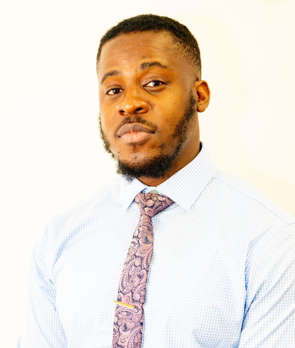 Professional wearing a light blue dress shirt and patterned pink tie stands confidently against a white background.