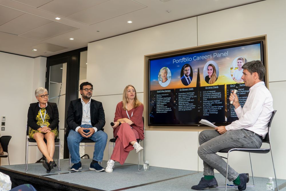 People sit around a modern white office space during a business meeting while others watch from a video call.
