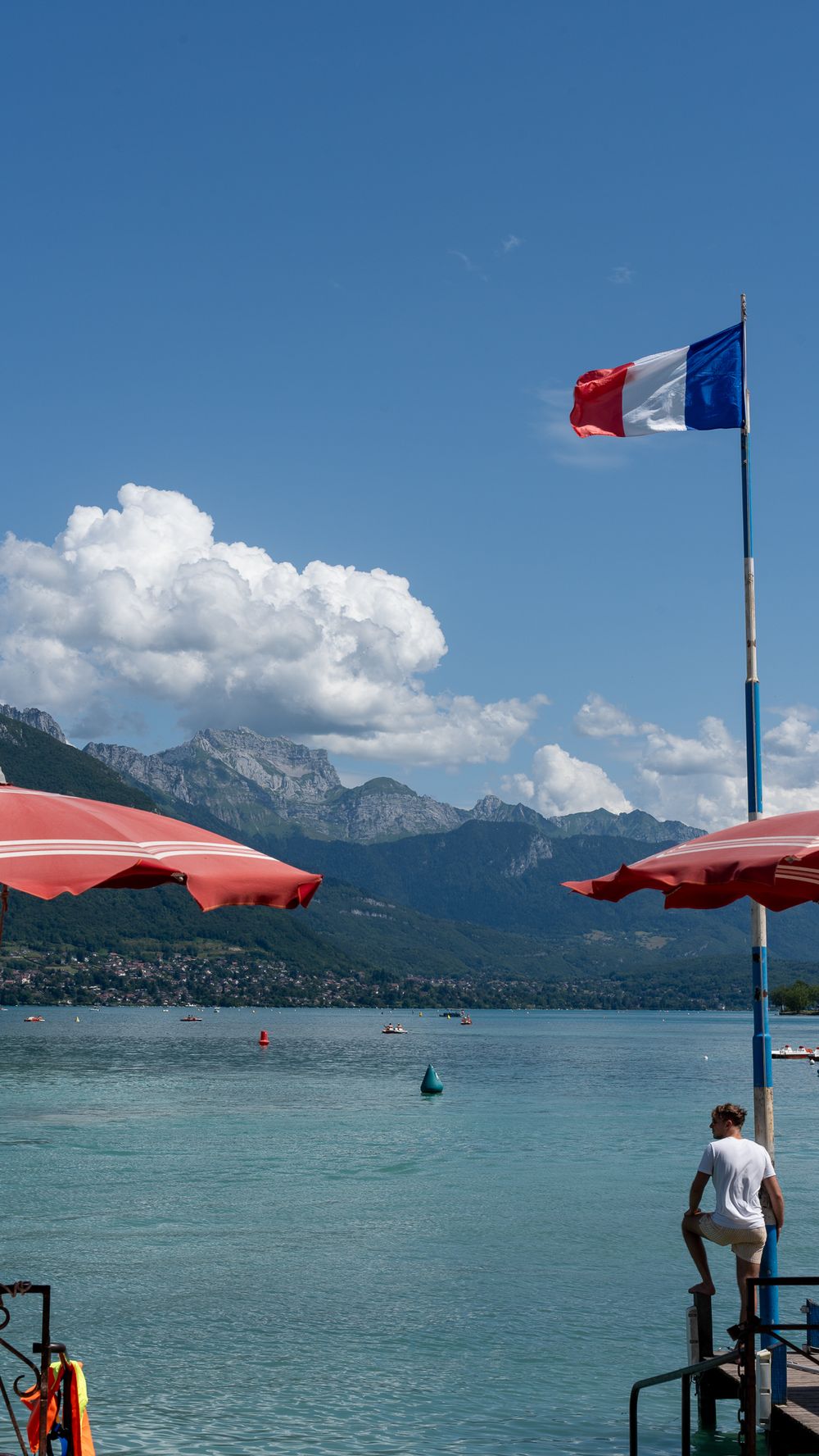 French flag waves over Lake Annecy with red umbrellas and mountains in background.