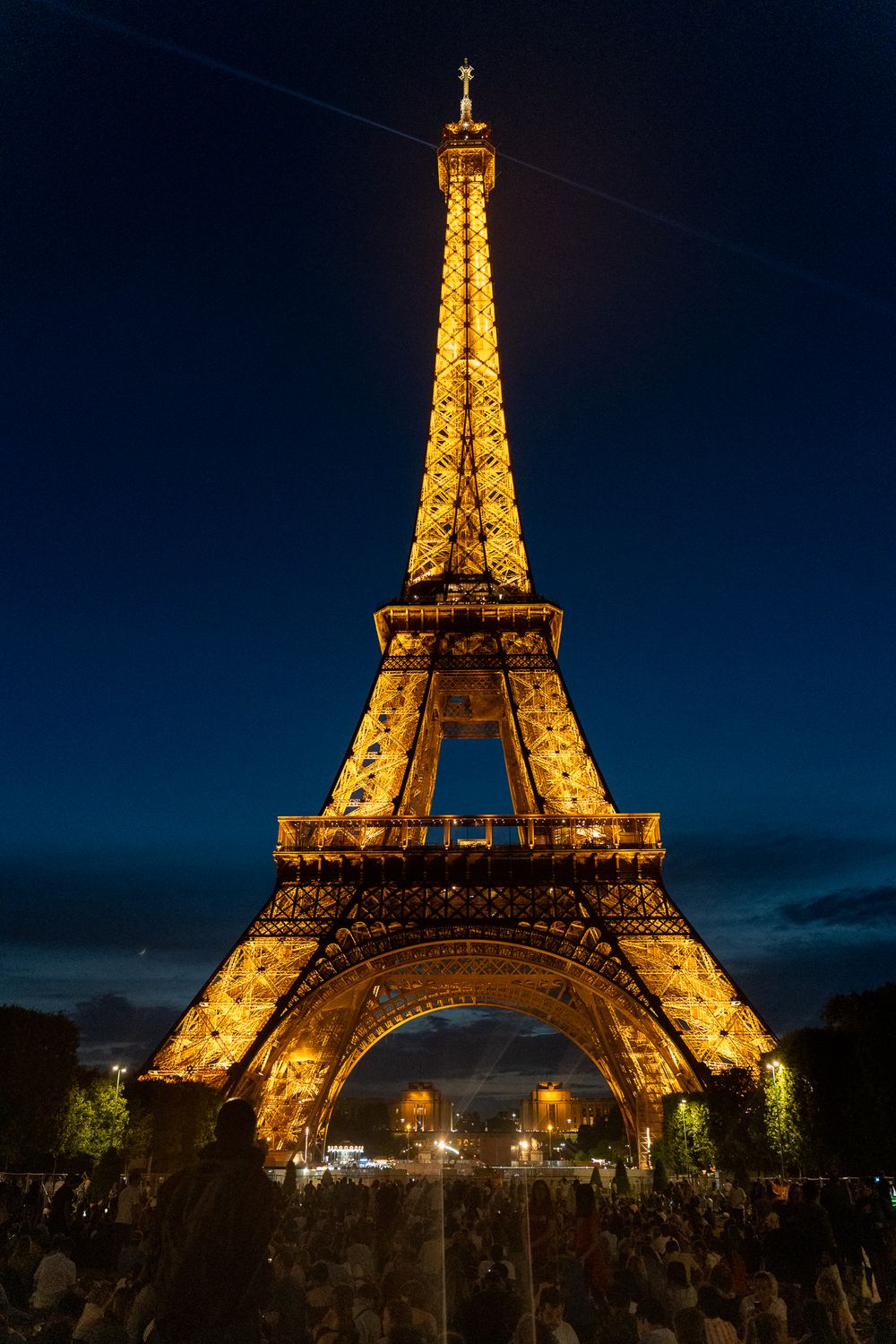 The illuminated Eiffel Tower glows golden against a dark blue night sky in Paris, with crowds gathered below.