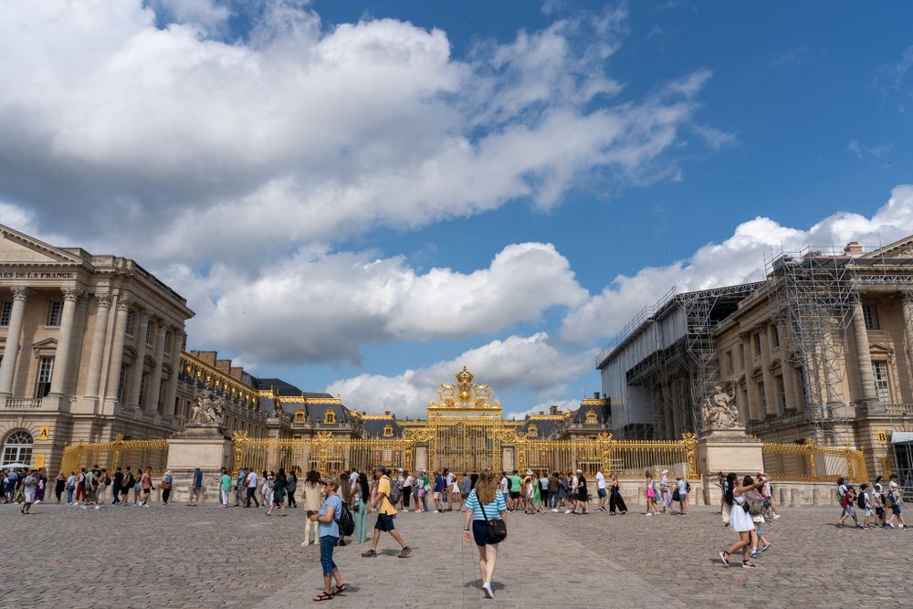 The grand Palace of Versailles courtyard with its ornate buildings under blue skies and white clouds.