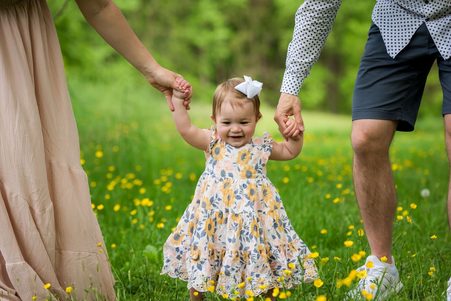 A toddler in a floral dress walks with parents among spring wildflowers.