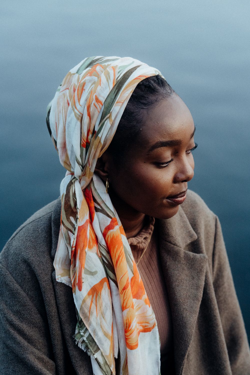 Portrait of person wearing patterned headscarf and coat against blue water background.