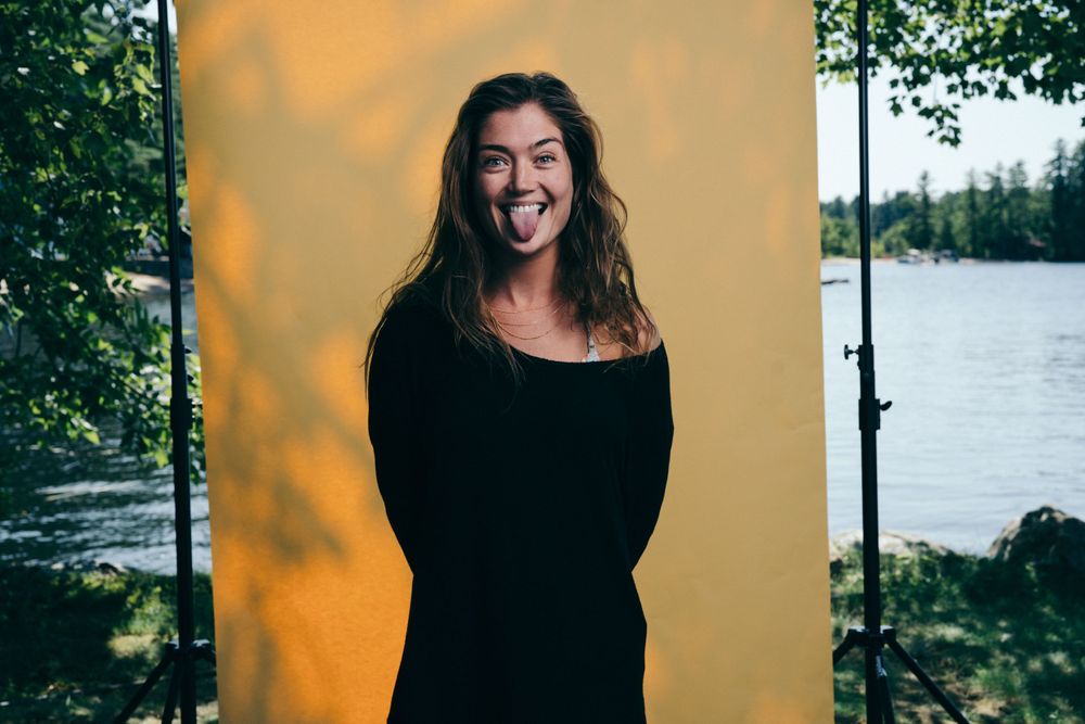 Someone in a black sweater stands outdoors by a lake sticking their tongue out playfully against a sunset-lit backdrop.