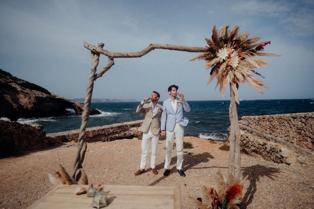 Two people celebrate under a rustic beach wedding arch with dried palm fronds.
