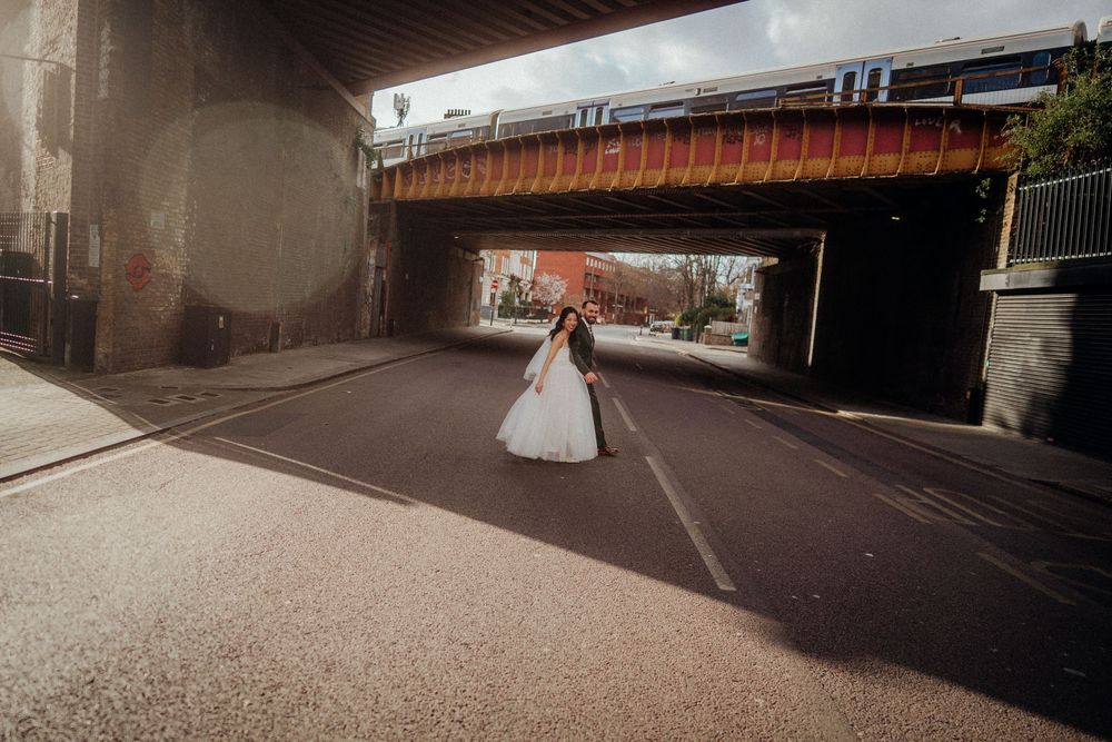 Bride in white gown stands in the middle of an urban road beneath a railway bridge on her wedding day.