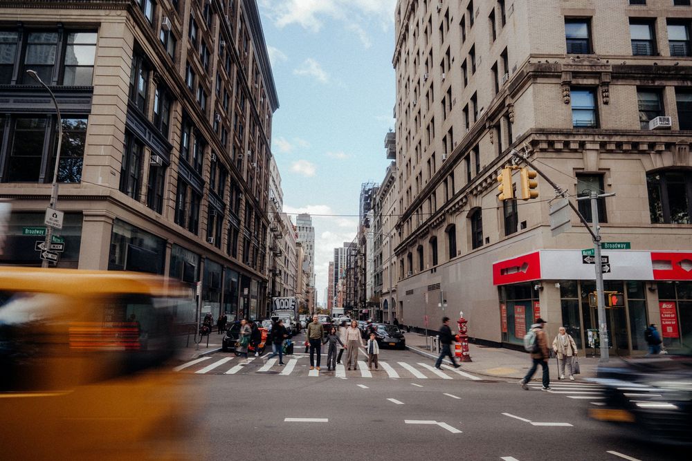 Busy New York City street intersection with yellow taxi, pedestrians crossing zebra crossing, and tall urban buildings.
