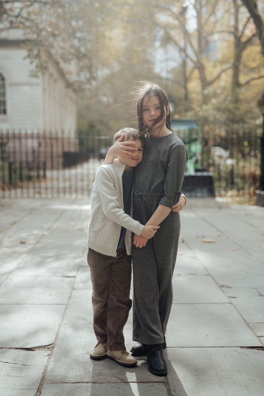 Two children sharing a warm hug on a city sidewalk surrounded by autumn trees.