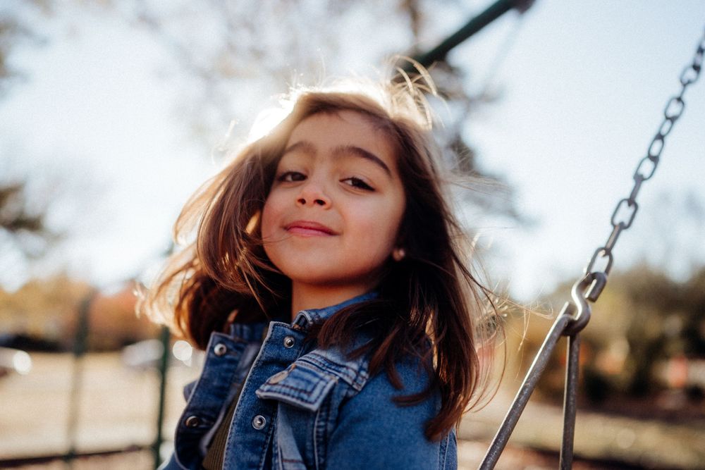 A young person on a swing smiles brightly in denim jacket against a blurred background.