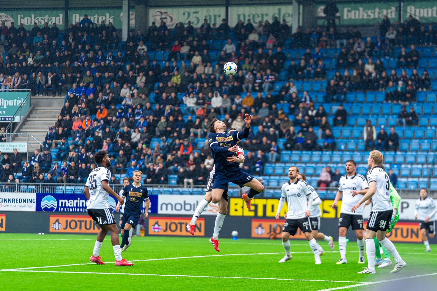 Soccer players compete for a header during a professional match in a packed stadium with advertising boards visible.