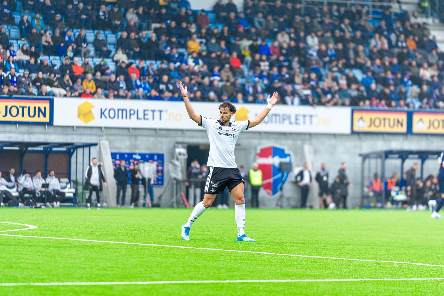 Soccer player in white uniform celebrates on green field with stadium crowd and Jotun and Komplett sponsor banners behind him.