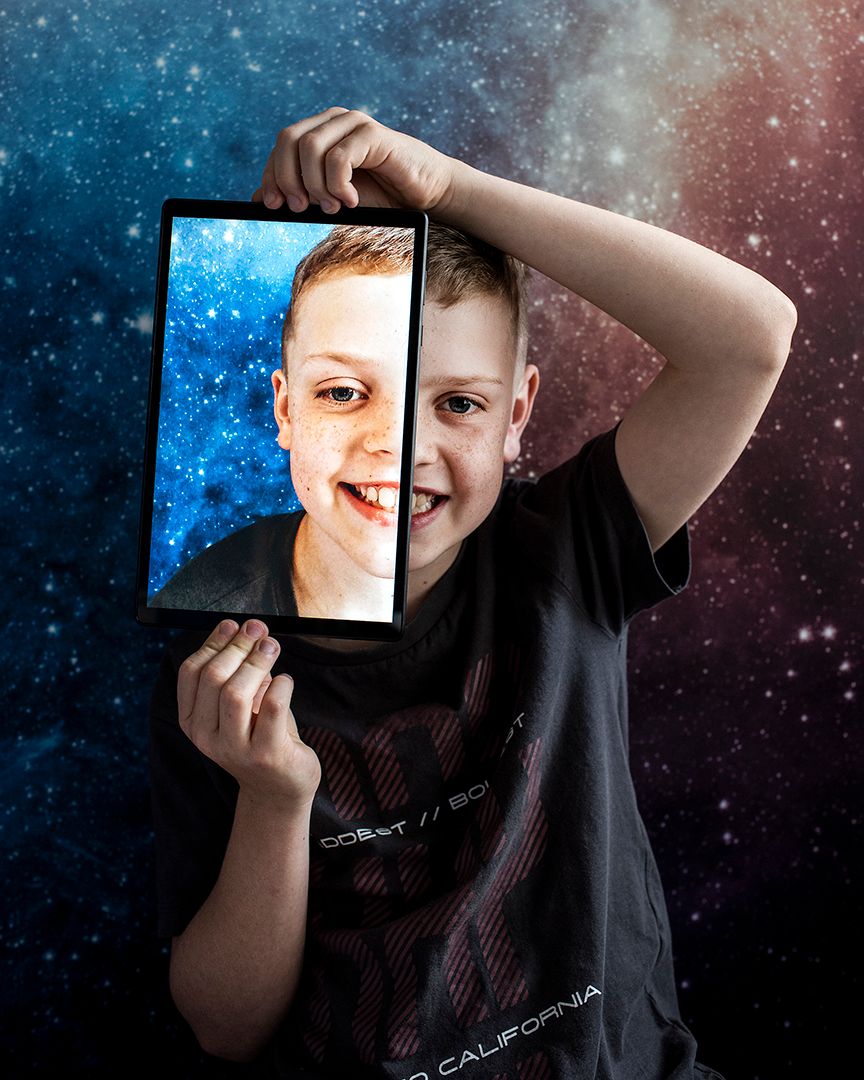 Portrait of a white boy, he is smiling holding an ipad up to his face. The ipad also shows a portrait of the boy's face