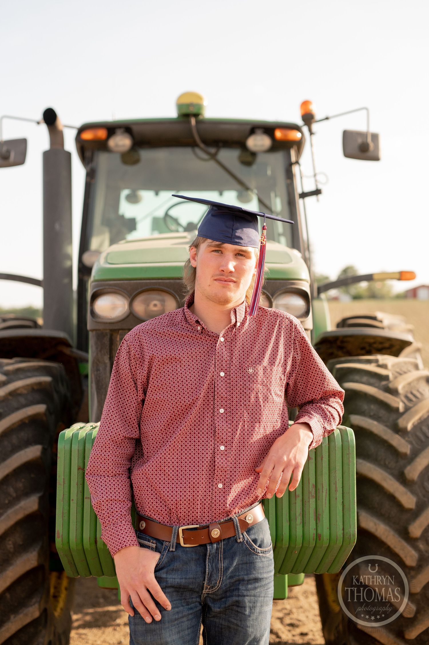 Graduate in plaid shirt poses with John Deere tractor in rural farm setting.
