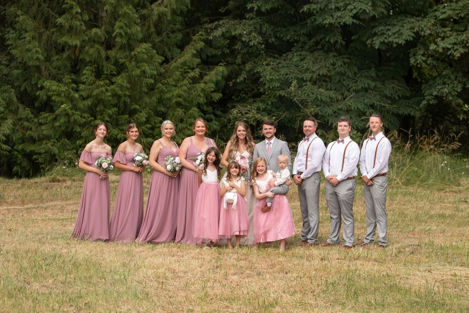 Wedding party in gray suits and coral dresses poses outdoors in a garden setting.