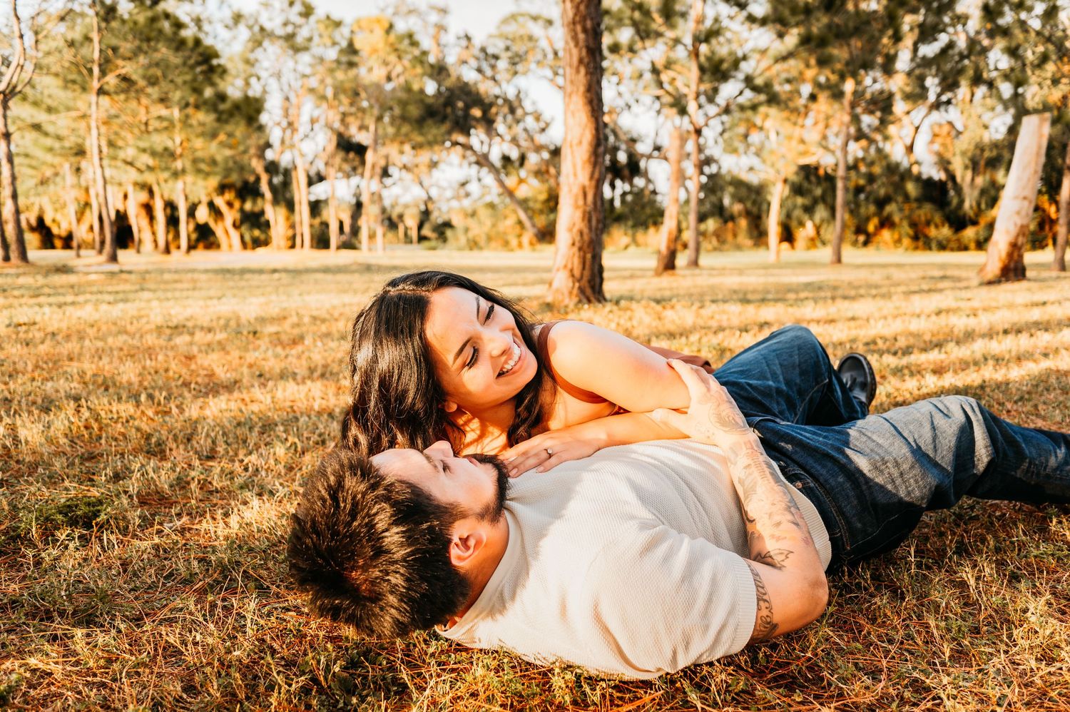 A couple laying together on autumn leaves in a golden-lit forest setting during sunset.