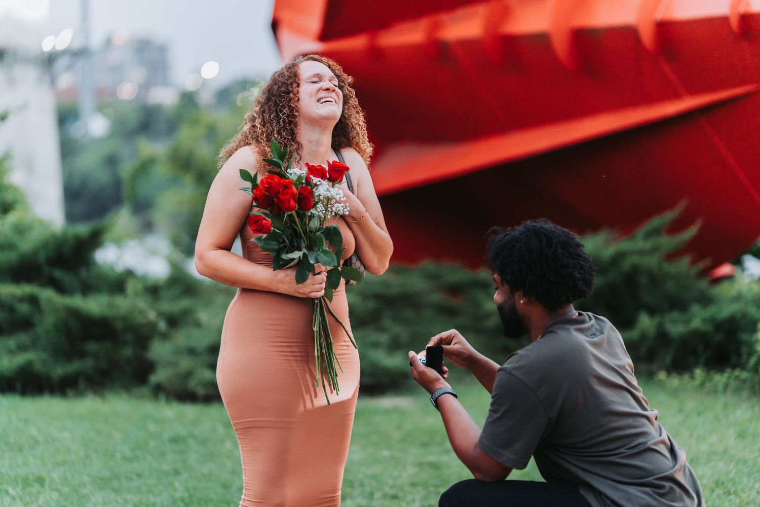 A romantic proposal moment outdoors with red roses and a peach-colored dress against a vibrant red architectural backdrop.