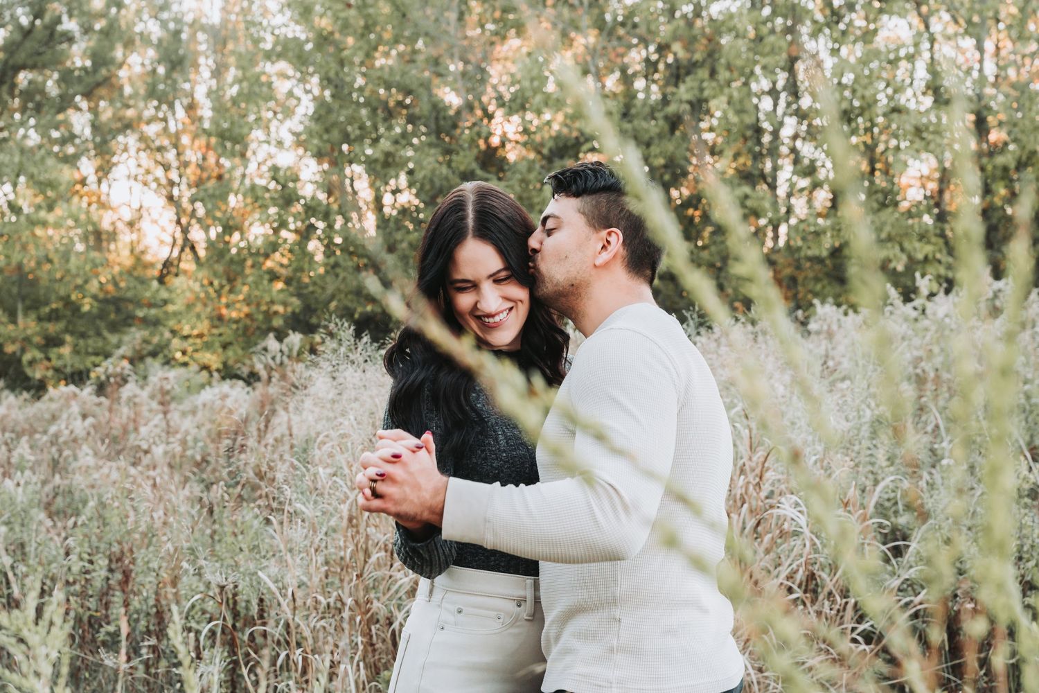 A couple embracing and laughing together in a natural outdoor setting with sunlit trees in the background.