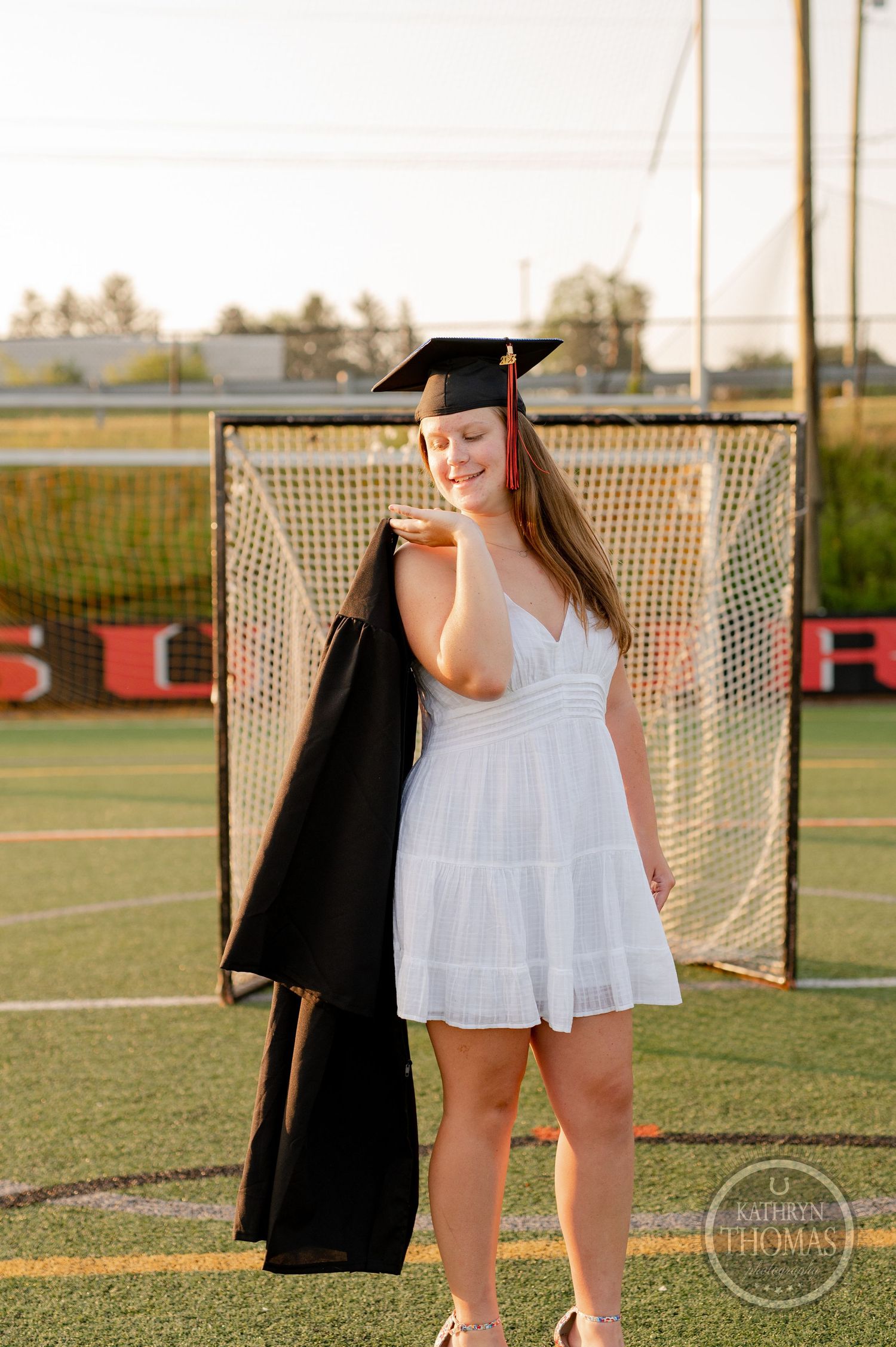 Graduate in white dress and graduation cap poses in front of lacrosse goal at sunset.