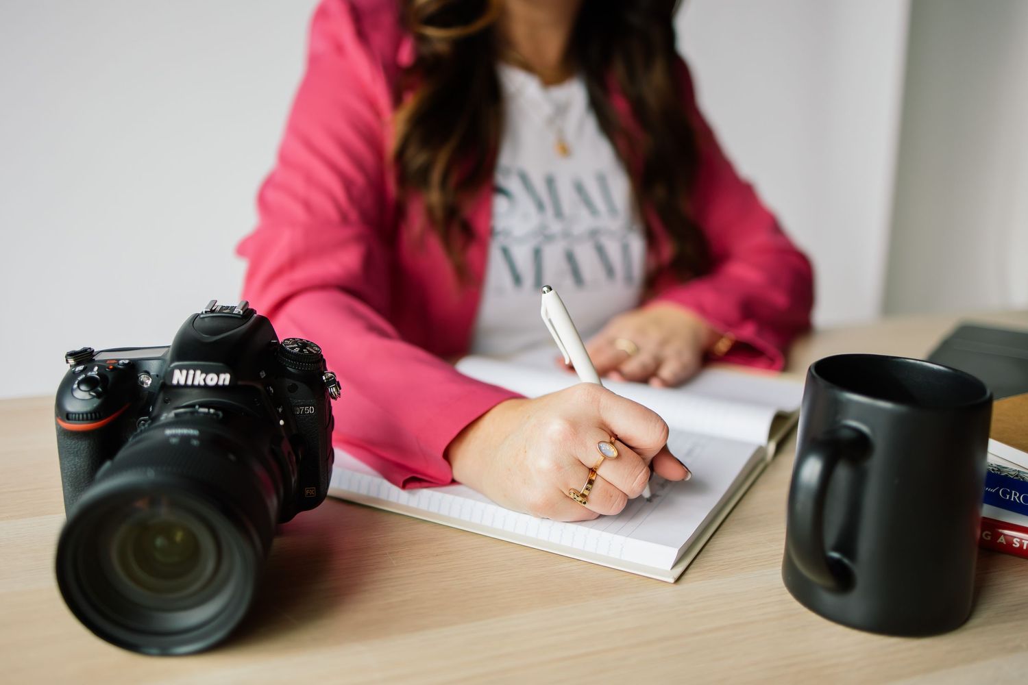 Woman in pink blazer writing in notebook beside Nikon camera and black mug on wooden desk.