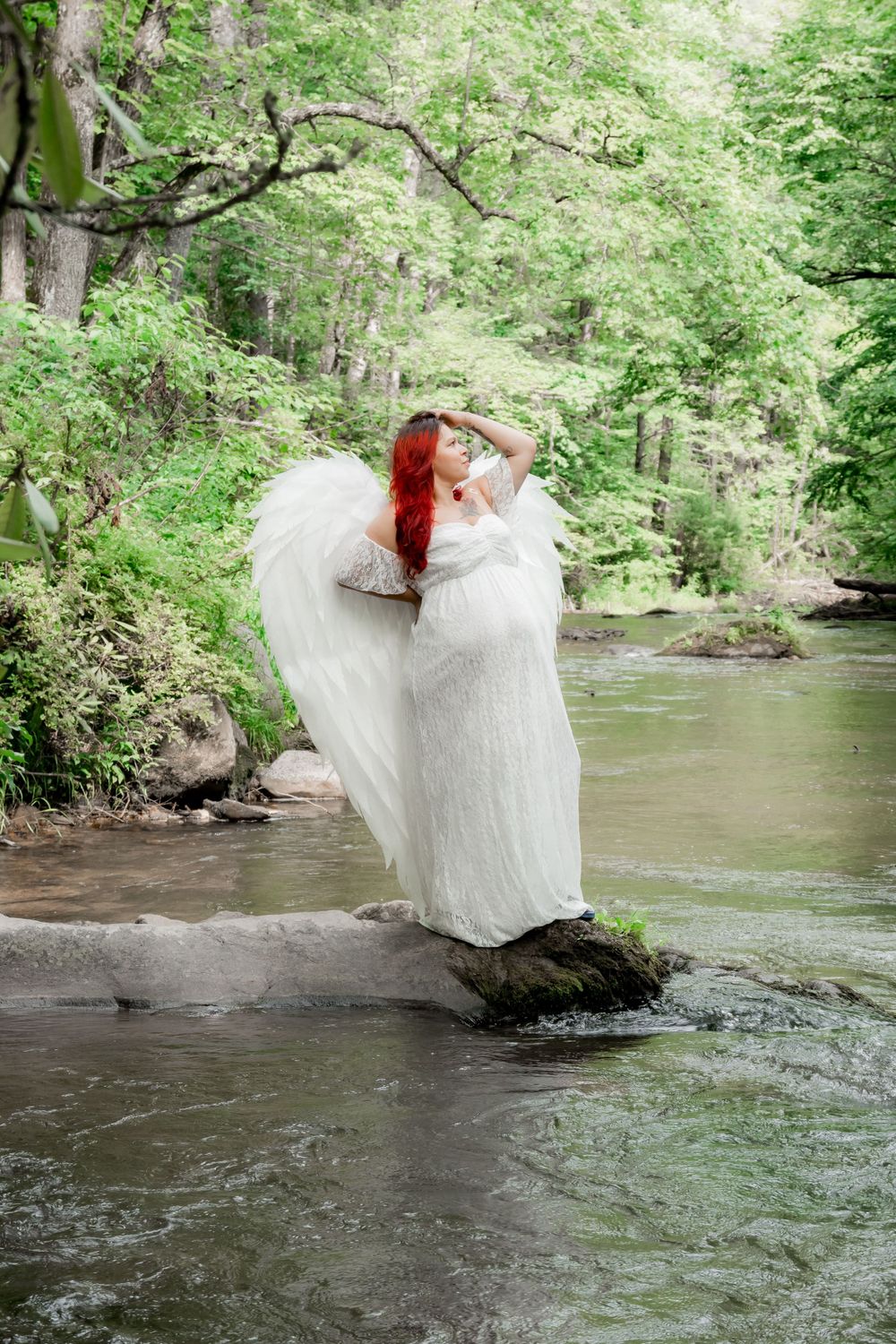Red-haired woman in white angel costume with wings stands on river rocks surrounded by lush green forest. Maternity Photographer.