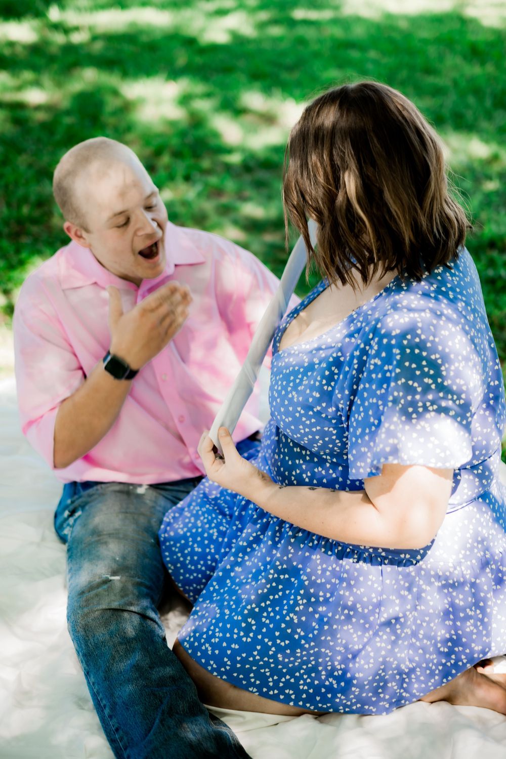 Excited man in pink shirt reacts to pregnant woman in blue polka dot dress during outdoor Pregnancy Announcement.