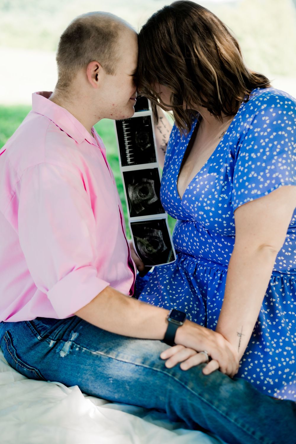 Couple sitting outdoors holding hands and an ultrasound photo, touching foreheads in a tender pregnancy announcement moment.
